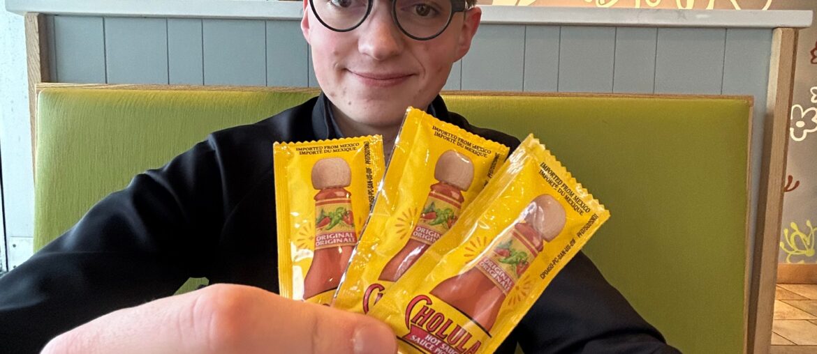 Brandon sitting in a booth at a restaurant holding up packets of hot sauce and smiling at the camera.