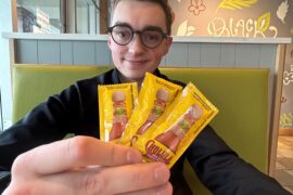 Brandon sitting in a booth at a restaurant holding up packets of hot sauce and smiling at the camera.