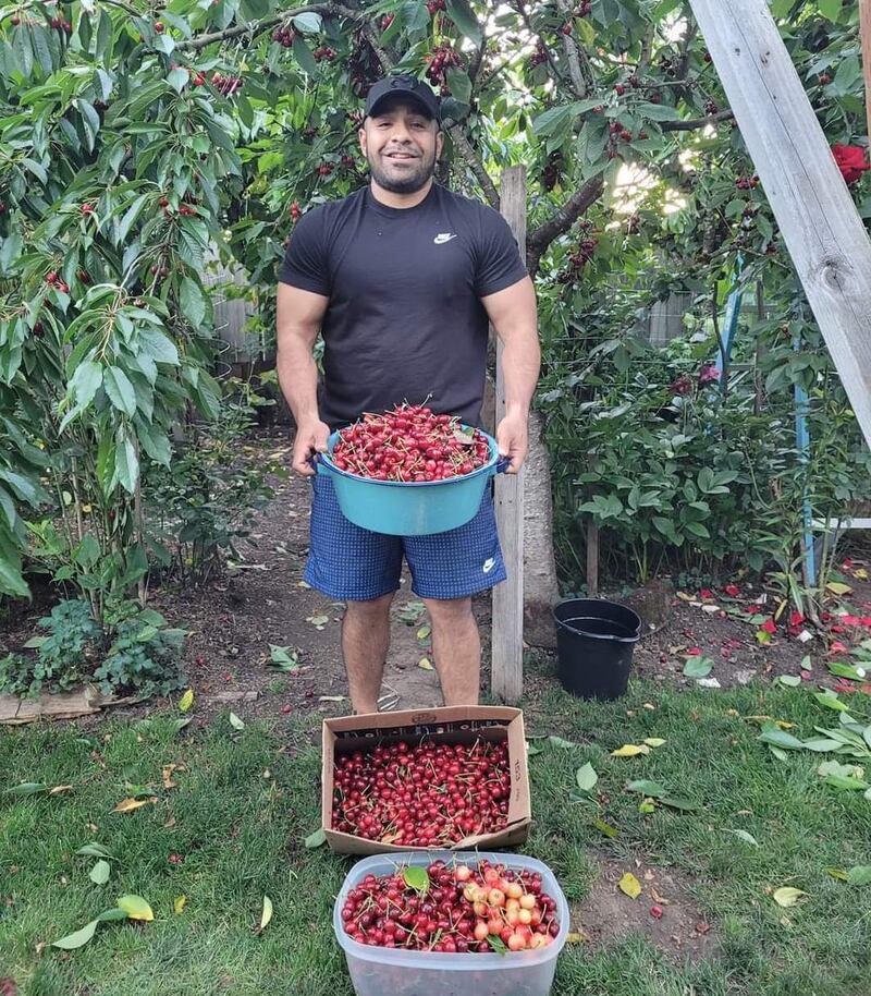 Ezequiel Lopez-Reyes poses in his yard, holding a pot full of cherries, with boxes of cherries at his feet.