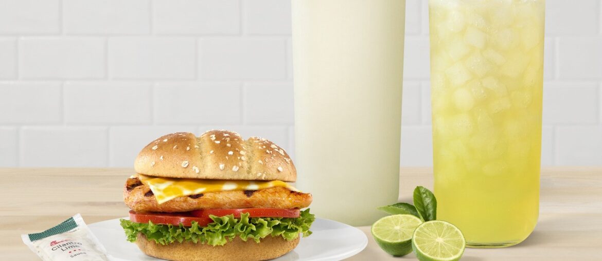 A Grilled Spicy Deluxe Sandwich on a plate with two Seasonal Key Lime Beverages, and limes on the side.