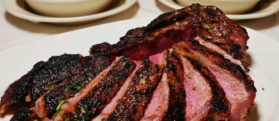Sliced barbecued ribs on a white plate, dark crusted exterior and pink smoke ring visible, with side dishes in the background.