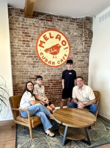 A family of four poses in front of a rustic brick wall inside Melao! Cuban Cafe. A woman and a young boy sit on a vintage-style tan leather armchair, while a man sits in a similar chair across from them. A second, older boy stands behind the first. Between them is a round wooden coffee table with black metal legs. On the brick wall above them hangs a large circular logo featuring a stylized orange bird and the words "MELAO! CUBAN CAFE"