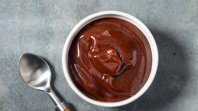 Small white ramekin of brown barbecue sauce next to silver spoon on gray surface
