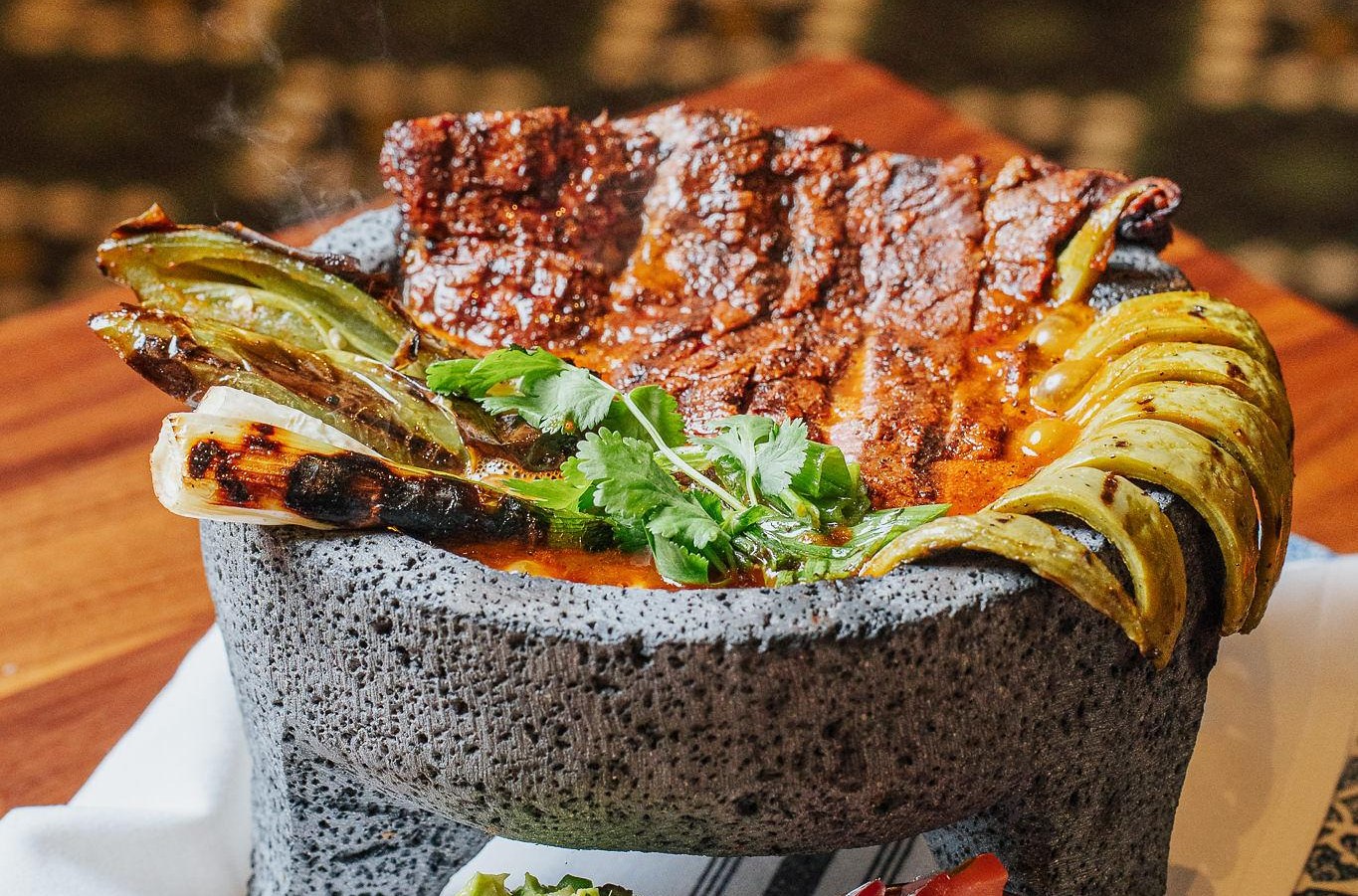 A stone bowl draped with grilled veggies, meat, nopales, and fresh cilantro with sides of salsa and guac.