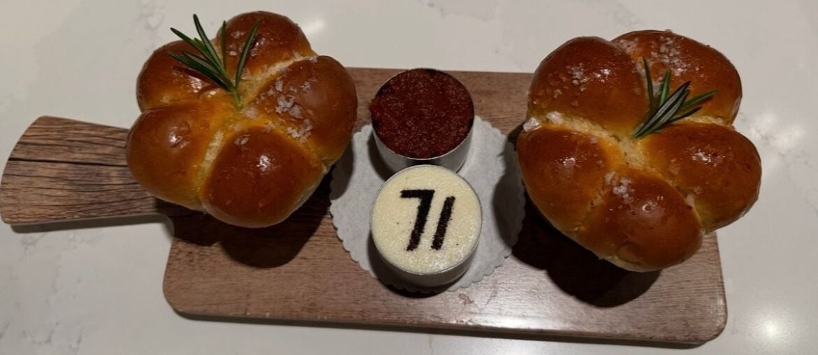 A delicious preview of Steakhouse 71 at Disney’s Contemporary Resort: two round rosemary-topped bread rolls and two mini desserts—one featuring the “71” logo—beautifully presented on a wooden board. Reservations are a must for this popular Walt Disney World dining spot.