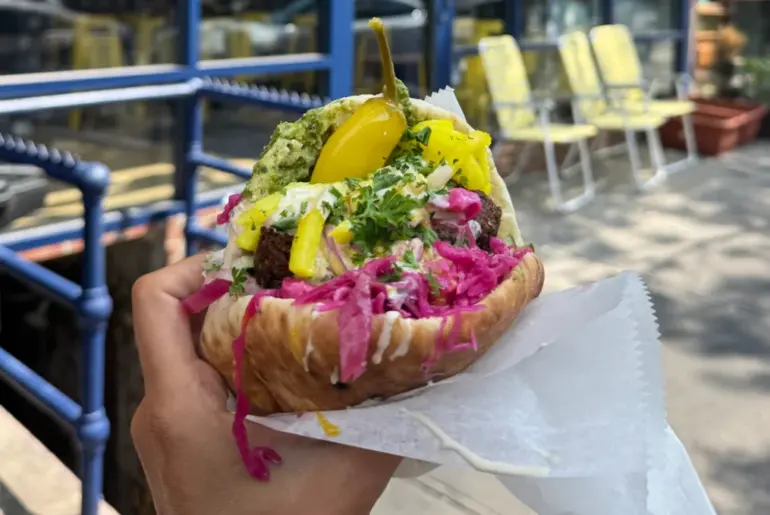 A hand holds a pita stuffed with falafel, yellow pickles, red cabbage, herbs, and creamy sauce. Outdoor seating with yellow chairs and tables is visible in the blurred background.