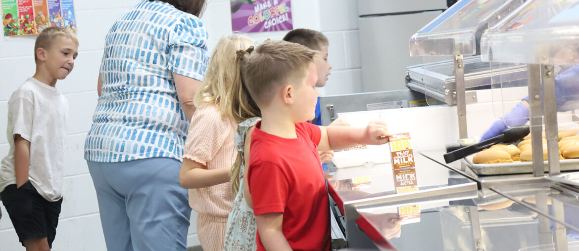 A group of young students stand in a cafeteria line waiting to pick up their lunches.