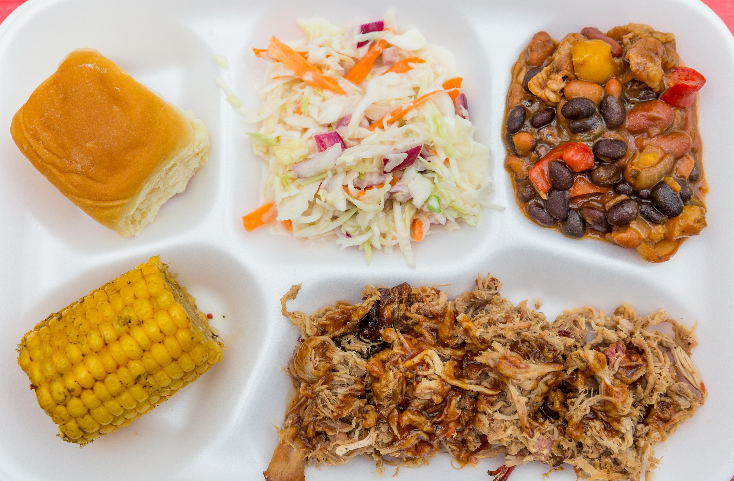 A styrofoam platter with pulled pork, corn, coleslaw, and beans.