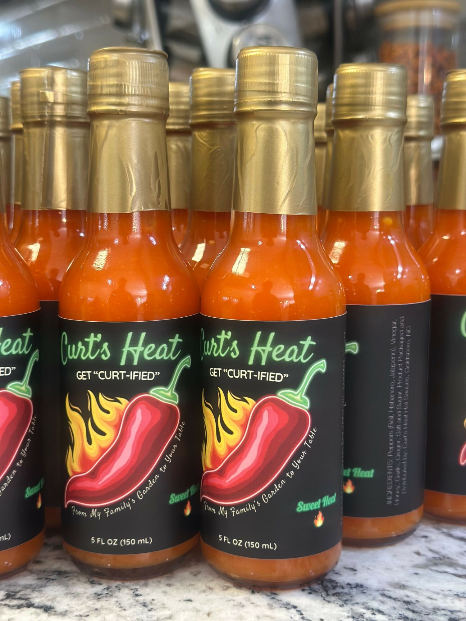 Multiple bottles of hot sauce with black labels and red chili pepper graphic, arranged on a surface.