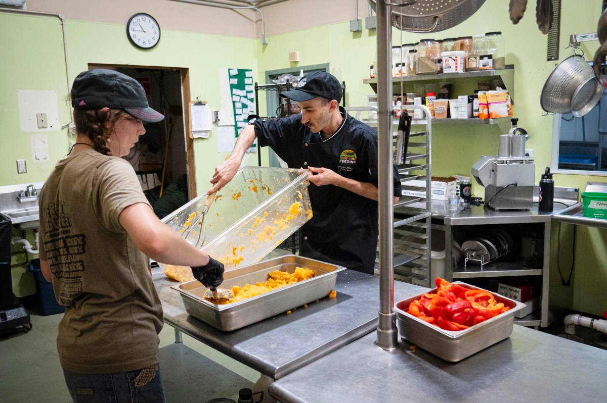 Two people prepare food in a commercial kitchen, transferring chopped vegetables from a large container into metal trays on a stainless steel counter.