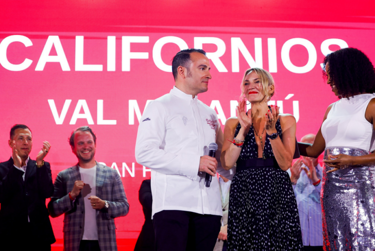 A man in a white chef jacket holds a microphone while a woman in a black dress claps, with people applauding behind them against a red background.