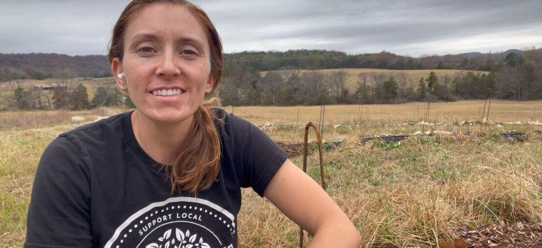 A person wearing a “Support Local Farms” T‑shirt sits in a cultivated field with rural farmland and rolling hills in the background, representing community‑based agriculture and local food access efforts.