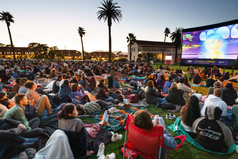 A large crowd sits on blankets and chairs on grass, watching an animated movie at an outdoor cinema during sunset, with palm trees and buildings in the background.