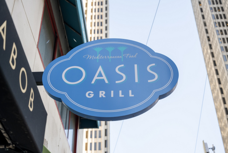 A blue oval sign with white and green text reading "Mediterranean Food OASIS GRILL" hangs outside a building in an urban area.